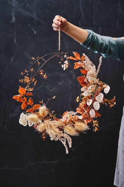 Floristería femenina irreconocible demostrando corona floral hecha de ramitas con hojas secas y plantas en tienda floral contra pared de mármol - Foto, Imagen