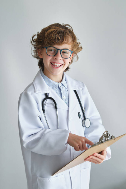 Criança alegre em roupão médico com estetoscópio mostrando prancheta enquanto olha para a câmera no fundo branco - Foto, Imagem