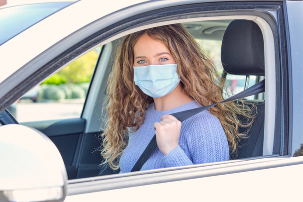 Giovane attraente conducente donna con maschera viso e maglione blu seduto nel sedile del conducente auto con cintura di sicurezza allacciata e guardando la fotocamera - Foto, immagini