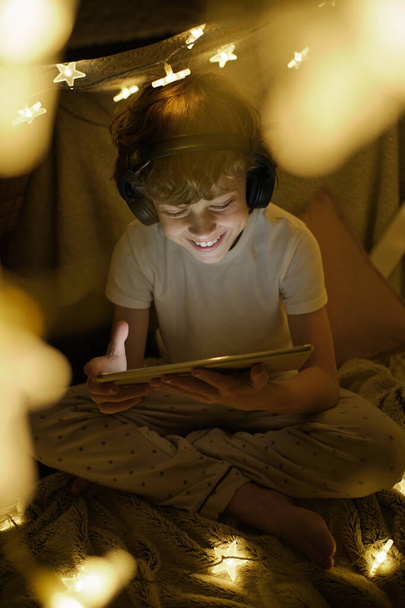 Cuerpo completo de chico positivo disfrutando de vídeo en la tableta sentado en la carpa manta decorado con guirnalda brillante - Foto, Imagen