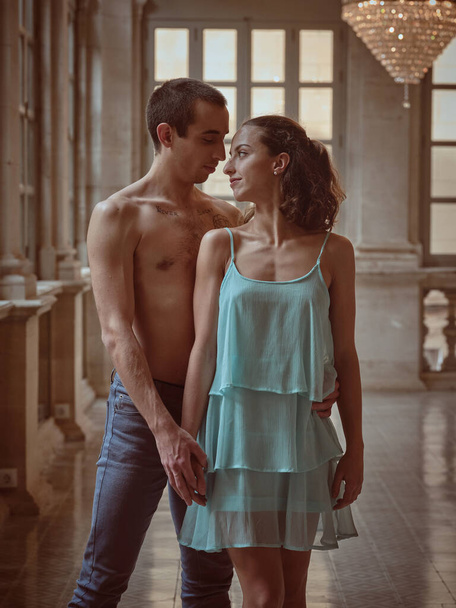 Loving shirtless guy in jeans and lady in blue dress and looking at each other while standing in palace interior - Photo, Image
