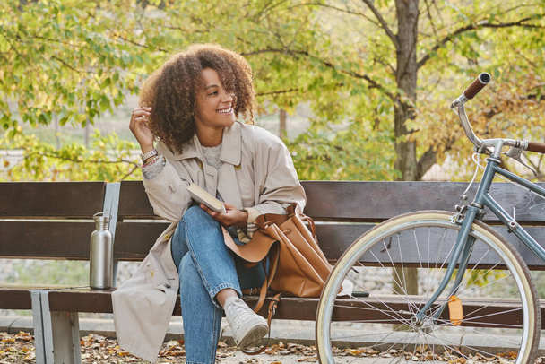 Pozitivní mladá dáma sedí s knihou v blízkosti kola na dřevěné lavičce při dotyku vlasů a odvracejí pohled - Fotografie, Obrázek
