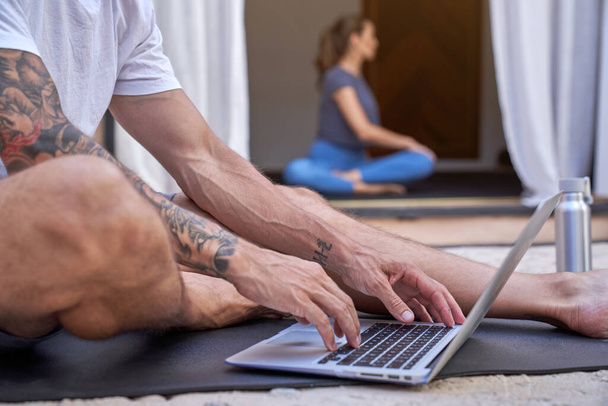 Cortar anônimo internet surf masculino no netbook enquanto mulher borrada praticando ioga no terraço durante o dia - Foto, Imagem