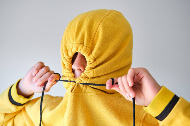 Enfant en sweat à capuche jaune serrant les lacets sur le capuche au-dessus de la tête révélant le nez sur fond gris - Photo, image