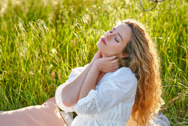 Atractiva joven hembra con el pelo ondulado con vestido blanco tocando el cuello suavemente mientras se sienta con los ojos cerrados en el césped abundante exuberante en el día soleado de verano - Foto, Imagen