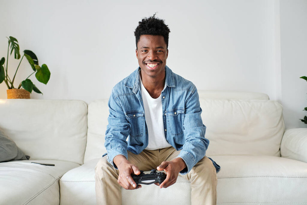 Wesoły młody Afroamerykanin w luźnym ubraniu siedzi na wygodnej kanapie z joystickiem i patrzy w kamerę z uśmiechem - Zdjęcie, obraz
