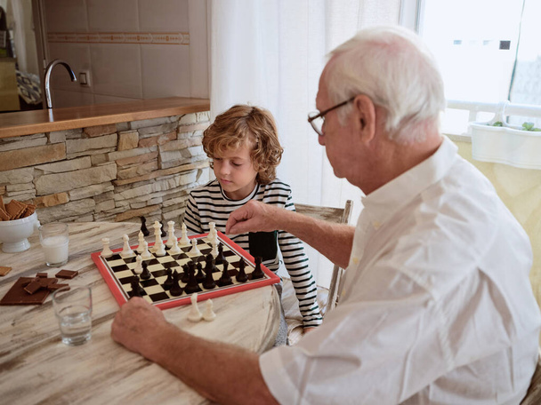 Vista lateral do homem idoso sentado à mesa de madeira com barra de chocolate e jogando xadrez com menino na cozinha - Foto, Imagem