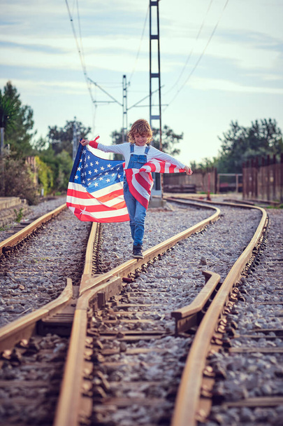 Kid segurando bandeira dos EUA com sorriso e andando ao longo da estrada de ferro perto interruptor sob céu azul nublado - Foto, Imagem