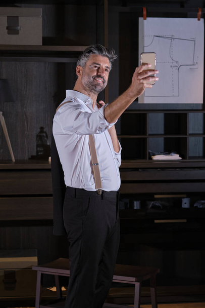 Позитивный предприниматель средних лет в костюме и подтяжках улыбается и делает автопортрет на смартфоне, стоя в гримерке - Фото, изображение