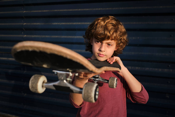 Chlapec s kudrnaté vlasy kontrolu skateboard proti tmavě modré zdi při tréninku na ulici - Fotografie, Obrázek