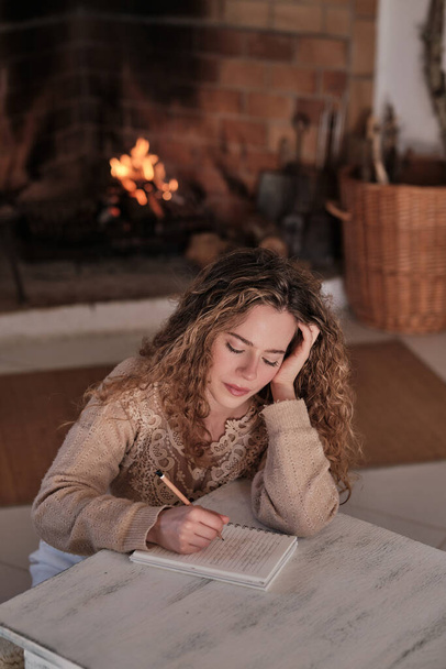 Inspirowana kobieta w ubraniu casual siedzi przy drewnianym stole w pobliżu płonącego kominka i robi notatki w notatniku w przytulnym salonie w domu - Zdjęcie, obraz