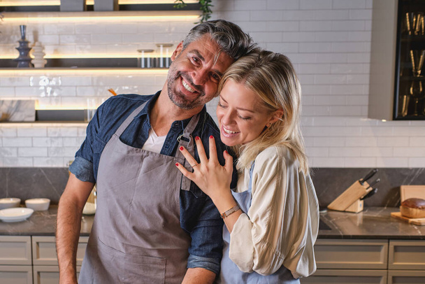 Позитивная женщина обнимает мужа в фартуке, смотрит в камеру с улыбкой во время приготовления пищи вместе на кухне рядом со стойкой с различными посудой - Фото, изображение