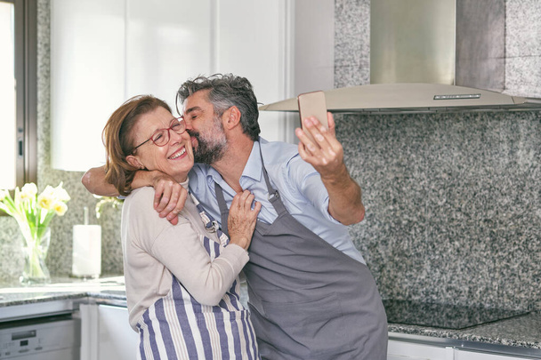 Feliz hombre maduro y mujer mayor positiva en delantales abrazándose y divirtiéndose mientras toma selfie en la cocina moderna - Foto, Imagen