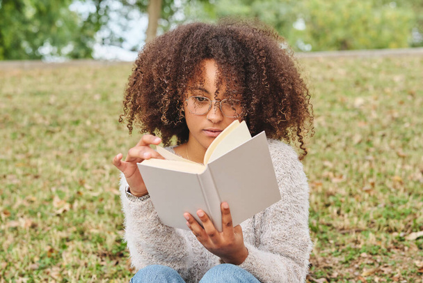 Zaostřená Afroameričanka s afro účesem sedí na trávě a čte zajímavý román v zeleném parku na rozmazaném pozadí - Fotografie, Obrázek