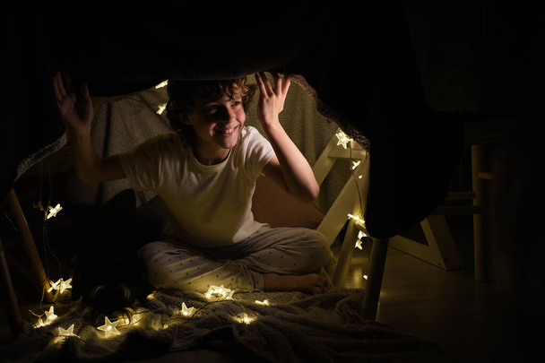Full body of cheerful boy looking away while sitting under blanket construction with glowing garland in dark room at home - Photo, Image