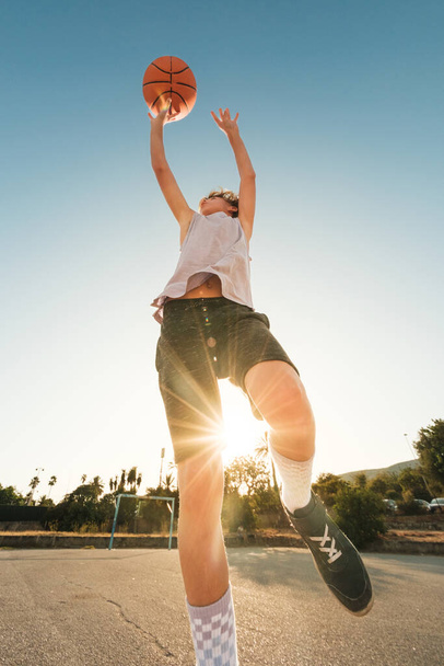 Bajo ángulo de niño en ropa deportiva lanzando bola naranja mientras juega streetball contra el sol en el campo de deportes - Foto, imagen