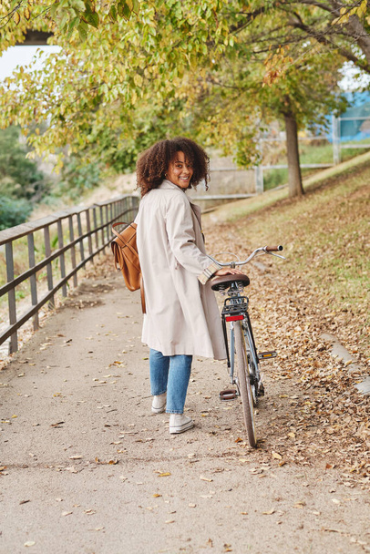 Vue arrière de la femme afro-américaine élégante avec un vélo à l'ancienne sur la piste dans le parc et en regardant la caméra - Photo, image