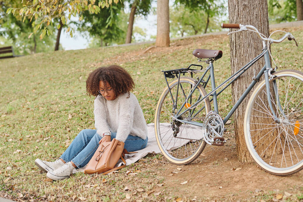 Cuerpo completo de mujer afroamericana sentada cerca de la bicicleta y buscando en la bolsa mientras descansa en el parque - Foto, imagen