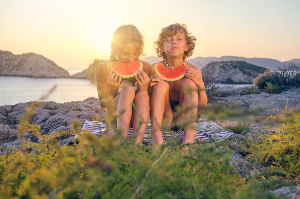 Corpo pieno di fratello e sorella positivo seduto su una coperta vicino al mare calmo sulla costa rocciosa e godendo di anguria fresca durante le vacanze estive - Foto, immagini
