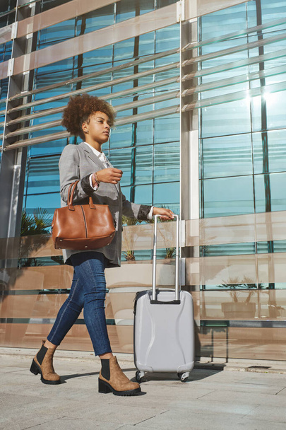 Full body of ethnic female with curly hair in smart casual wear smiling while standing with suitcase in city and talking on mobile phone - Fotó, kép