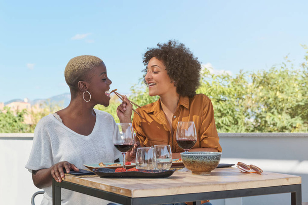 Smiling multiethnic women feeding each other Asian food while having lunch with red wine on terrace of restaurant - Photo, Image
