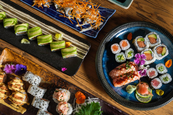 Верхний вид вкусных суши-роллов с рыбой и различной начинкой подается на подносе с декоративными цветами и васаби с имбирем - Фото, изображение