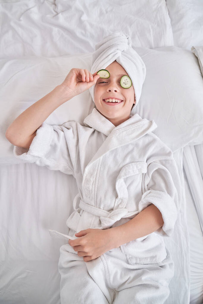 High angle of smiling child wearing towel turban demonstrating cucumber slice while resting on white bed during spa procedures in bedroom at home - Photo, Image