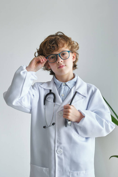 Bambino intelligente in vestaglia medica con stetoscopio e occhiali che guarda la fotocamera su sfondo bianco - Foto, immagini