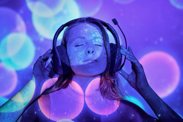 Mujer joven ajustando auriculares y disfrutando de buena música bajo la proyección de destellos de colores - Foto, imagen