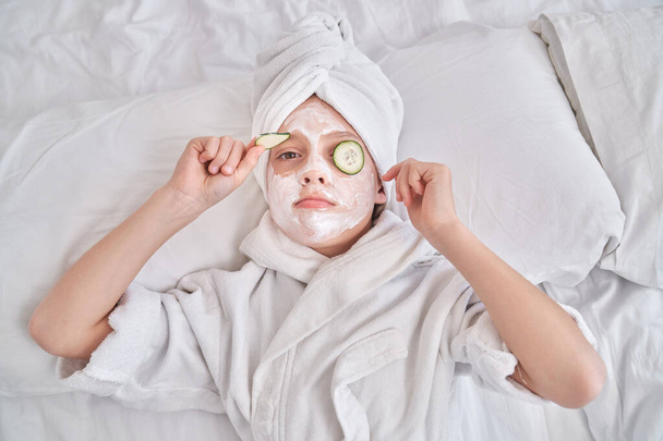 Top view of serious child in towel turban and bathrobe putting off cucumber slice and looking at camera while lying on bed during spa procedures in bedroom at home - Photo, Image