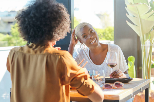 楽観的なアフリカ系アメリカ人女性がレストランのテーブルで友人の前に座って笑う - 写真・画像