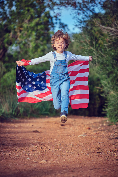 Pełne ciało energicznego chłopca z kolorowym machającym amerykańską flagą biegnącego szybko po wiejskich drogach w letni dzień - Zdjęcie, obraz