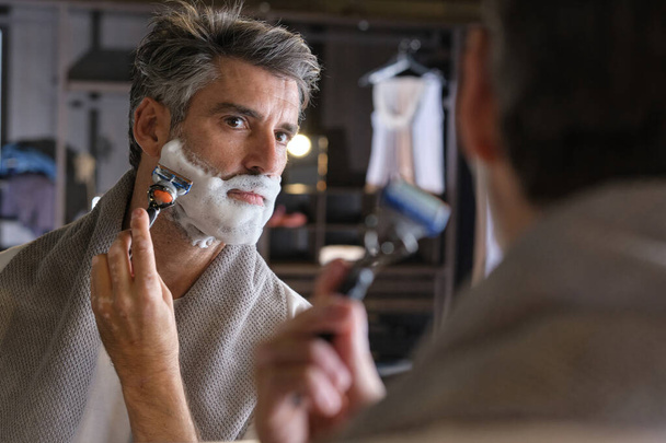 Adult gray haired man with foam on face shaving chin with razor near mirror at home - Photo, Image
