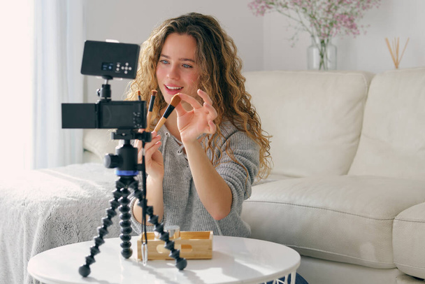 Młoda kobieta vlogger z długimi falowanymi włosami pokazując zestaw szczotek do makijażu twarzy na kamerze w salonie w domu - Zdjęcie, obraz