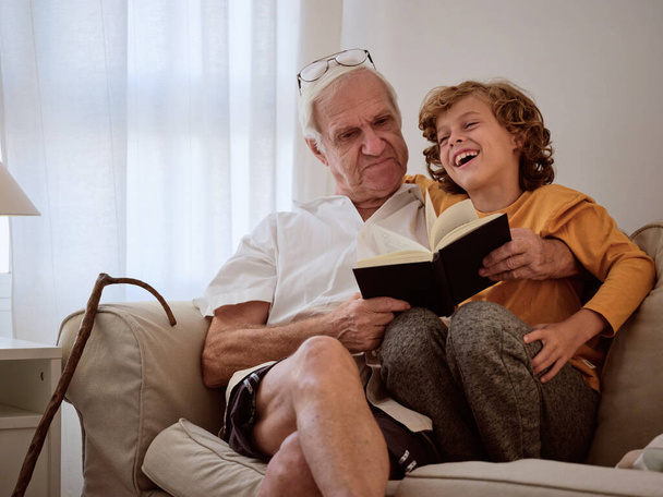 Niño sonriente abrazando anciano sosteniendo libro y sentado en un sofá acogedor en el día libre en la sala de estar - Foto, Imagen