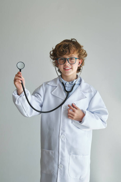 Симпатичный ребенок в медицинской форме и очках со стетоскопом, смотрящий в камеру на белом фоне - Фото, изображение