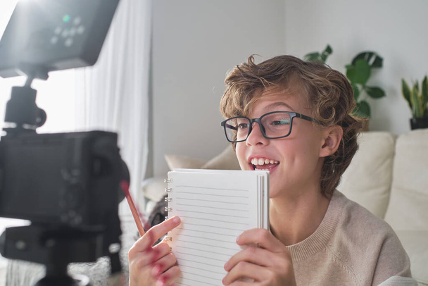 Cheerful preteen kid with curly hair looking at screen of camera and shooting video for blog while pointing at notebook and sitting in living room - Photo, Image