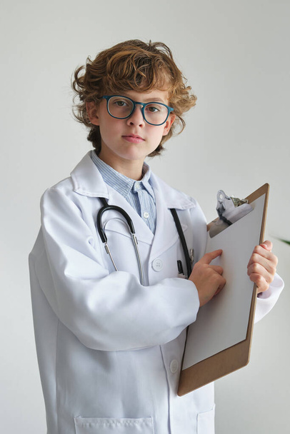 Smart child in eyewear and medical uniform pointing at empty paper on clipboard while looking at camera on white background - Photo, Image