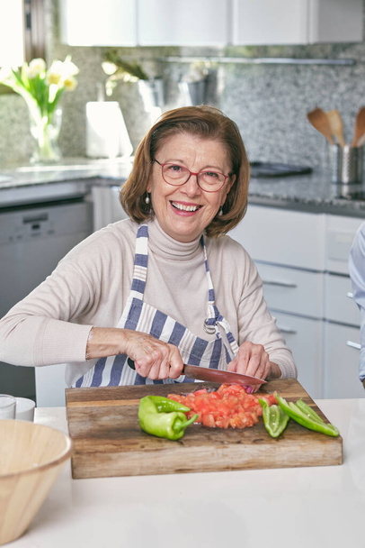 Donna anziana positiva in grembiule guardando la fotocamera con sorriso mentre tagliava pomodori freschi sul tagliere di legno in cucina - Foto, immagini