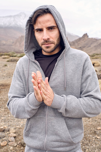 Adult serious bearded Hispanic male traveler in gray hoodie standing in mountains with clasped hands in Namaste gesture and looking at camera in Tenerife Canary Island Spain - Photo, Image