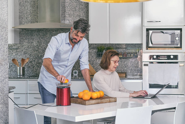 baard man in shirt het bereiden van gezonde sinaasappelsap in squeezer terwijl oudere vrouw in glazen browsen laptop in de keuken thuis - Foto, afbeelding