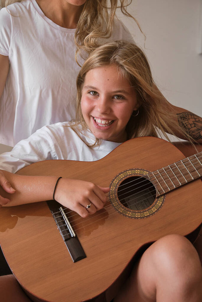 Függőleges fénykép egy szőke lány portréjáról, aki gitározik, miközben mosolyog a kamera előtt egy nővel, aki vele ül egy kanapén egy szobában. - Fotó, kép