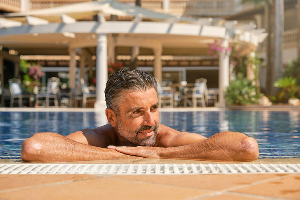 Cheerful bearded middle aged male tourist leaning on edge of swimming pool while resting in clean blue water during summer vacation in hotel - Photo, image