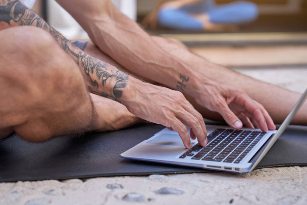 Anonymous tattooed male sitting on mat and typing on laptop keyboard during workout in gym - Photo, Image