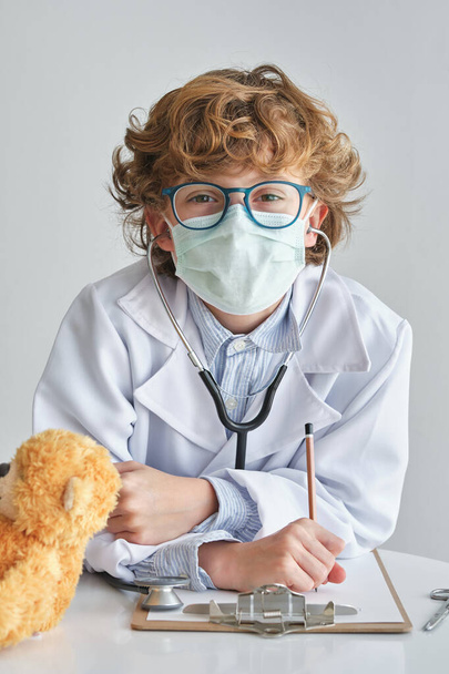 Ένα παιδί με αυτοπεποίθηση με ιατρική στολή και γυαλιά που κοιτάει την κάμερα. Ακούμπησε στο τραπέζι με μαλακή αρκούδα και πρόχειρο σε λευκό φόντο - Φωτογραφία, εικόνα