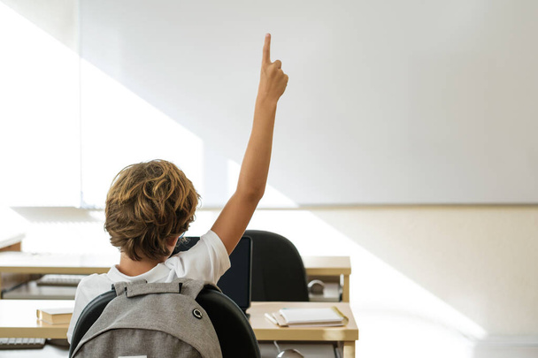 Widok z tyłu nierozpoznawalny chłopiec z podniesionym ramieniem siedzi przy stole w klasie podczas lekcji - Zdjęcie, obraz
