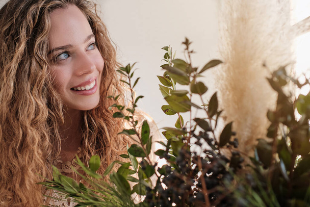 Positive blue eyed lady with curly blond hair looking away near green plants with fresh leaves - Photo, Image