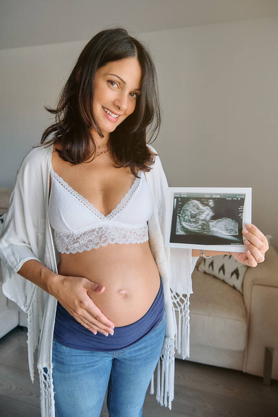 Восхитительная беременная женщина с обнаженным животом смотрит в камеру, стоя дома и демонстрируя ультразвуковое изображение ребенка - Фото, изображение