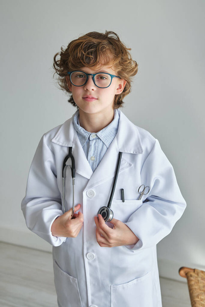 Умный ребенок в очках и медицинском халате с профессиональным инструментом, смотрящим в камеру - Фото, изображение