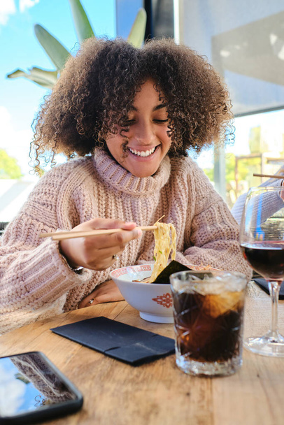 Содержание молодой афроамериканской женщины с вьющимися волосами в свитере, сидящей за деревянным столом и поедающей рамен палочками для еды - Фото, изображение